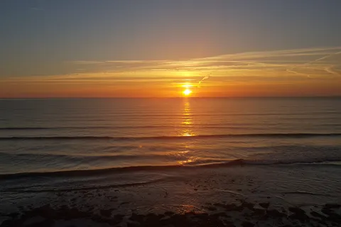 A low golden sun casts a bright reflection across the sea above small waves and a dark shoreline.
