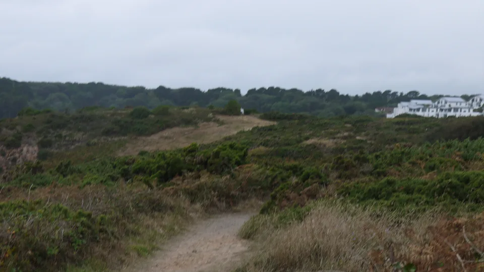 Rough grassland and scrub extend towards trees and distant white buildings beneath a cloudy sky.