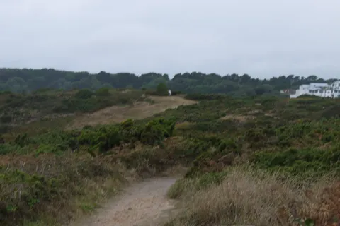 Rough grassland and scrub extend towards trees and distant white buildings beneath a cloudy sky.