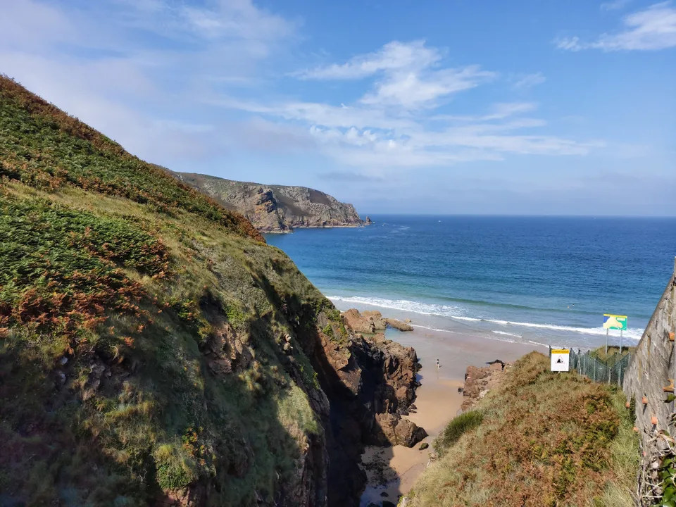 A steep grassy cliff overlooks the sandy shore and blue water at Plémont beneath scattered clouds.