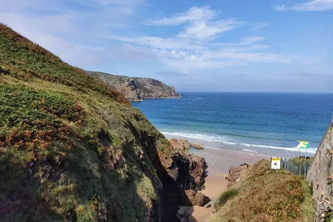 A steep grassy cliff overlooks the sandy shore and blue water at Plémont beneath scattered clouds.