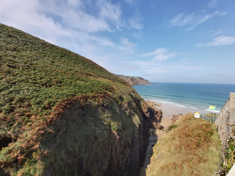 A green headland and rocky cliff edge frame a small sandy cove and blue sea at Plémont.