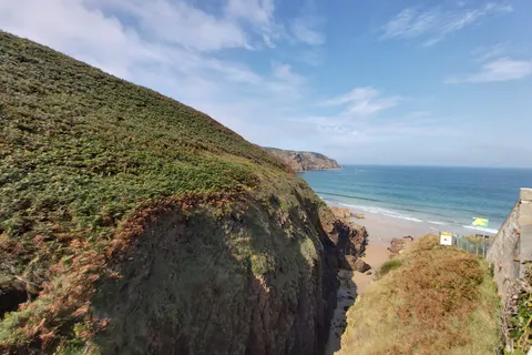 A green headland and rocky cliff edge frame a small sandy cove and blue sea at Plémont.