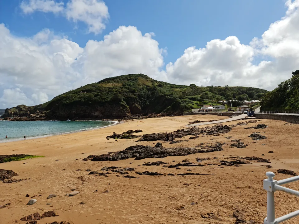 Golden sand and low dark rocks curve towards blue water and a green headland at Grève de Lecq.