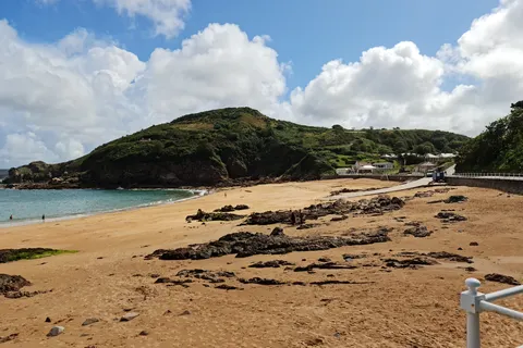 Golden sand and low dark rocks curve towards blue water and a green headland at Grève de Lecq.