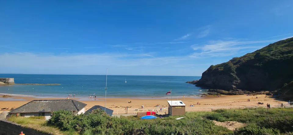 Low beach buildings and boats sit beside golden sand and blue water beneath a green headland at Grève de Lecq.