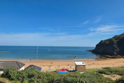 Low beach buildings and boats sit beside golden sand and blue water beneath a green headland at Grève de Lecq.