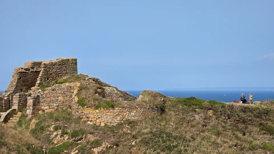 Grosnez Castle's stone ruins rise from a grassy slope above the blue sea with a clear sky overhead.