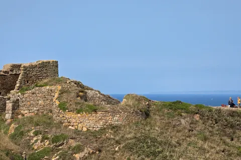 Grosnez Castle's stone ruins rise from a grassy slope above the blue sea with a clear sky overhead.