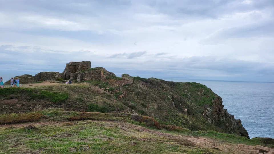 Low stone castle ruins stand on a rugged grassy headland above the sea at Grosnez beneath a clouded sky.