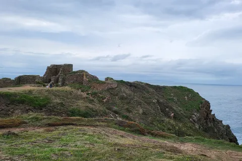 Low stone castle ruins stand on a rugged grassy headland above the sea at Grosnez beneath a clouded sky.
