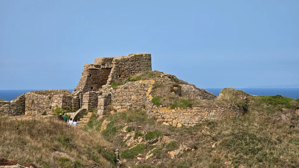 Rough stone castle walls rise above a grassy slope at Grosnez under clear blue sky beside the sea.
