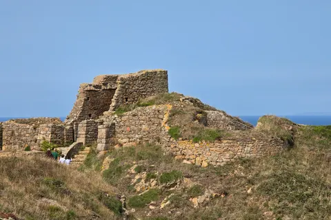 Rough stone castle walls rise above a grassy slope at Grosnez under clear blue sky beside the sea.