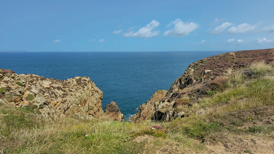 A rocky gap between grassy slopes opens to the blue sea at Grosnez beneath a clear sky.