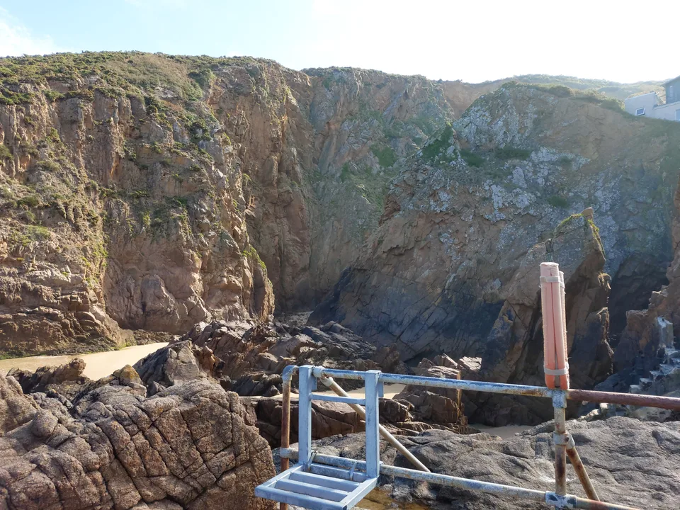 A metal handrail crosses rugged coastal rocks beneath a high cliff at Plémont, beside a shaded rocky recess.