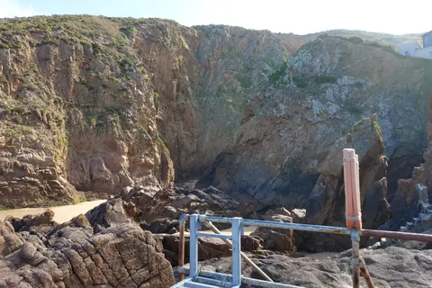 A metal handrail crosses rugged coastal rocks beneath a high cliff at Plémont, beside a shaded rocky recess.