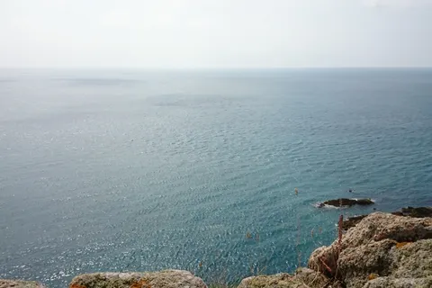 A calm blue sea meets a pale hazy sky beyond a rocky coastal edge with grass and lichen.