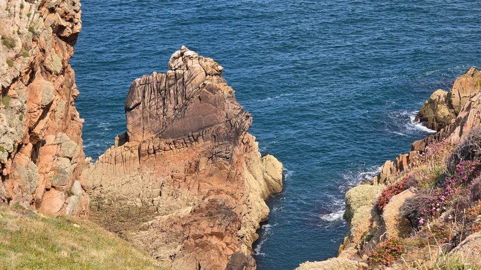 A tall ridged rock formation rises beside coastal cliffs over deep blue water at Grosnez.