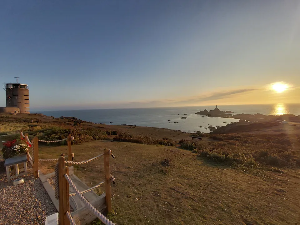 A coastal tower stands beyond picnic tables and a rope fence, with a golden sunset over the rocky sea beyond.