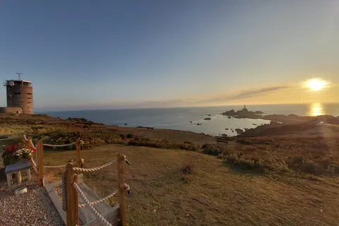 A coastal tower stands beyond picnic tables and a rope fence, with a golden sunset over the rocky sea beyond.