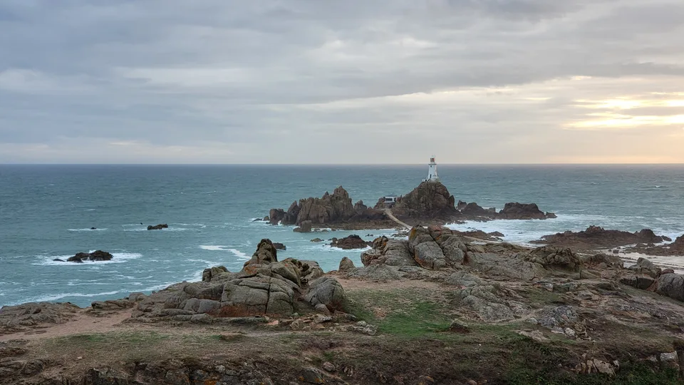 Dark rocks and turquoise water surround La Corbière Lighthouse beneath an overcast evening sky.