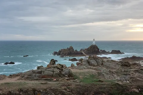 Dark rocks and turquoise water surround La Corbière Lighthouse beneath an overcast evening sky.