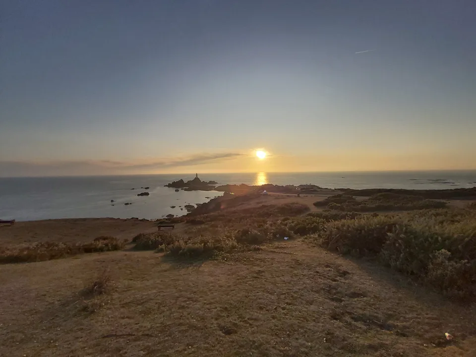 La Corbière Lighthouse stands on dark coastal rocks beside a bright reflection from the setting sun.
