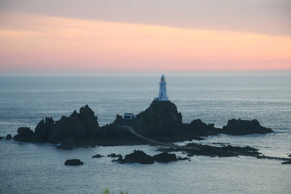 La Corbière Lighthouse stands on dark rocks against blue-grey water beneath a pink and peach evening sky.