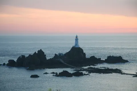 La Corbière Lighthouse stands on dark rocks against blue-grey water beneath a pink and peach evening sky.