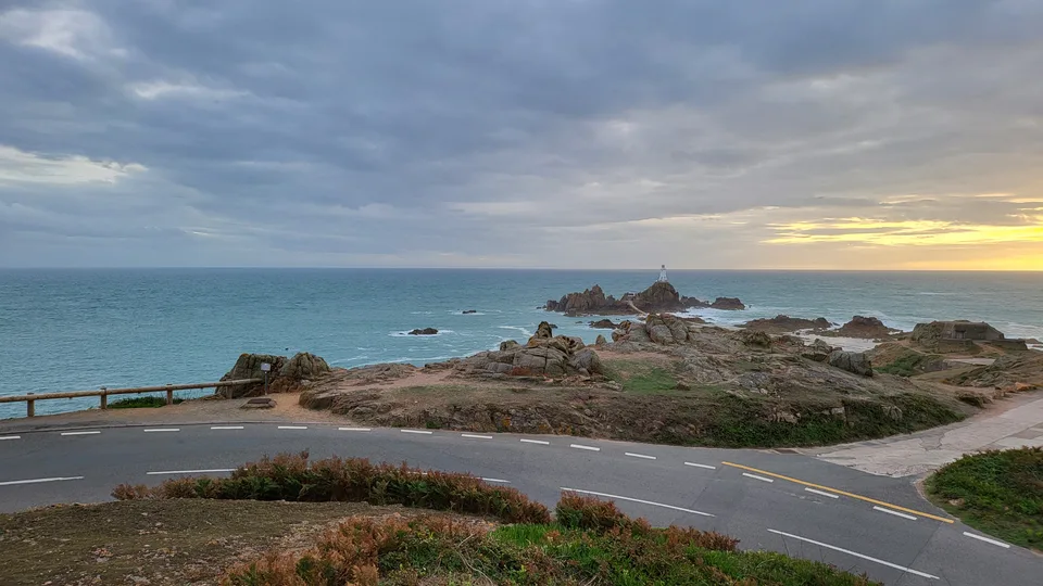 La Corbière Lighthouse stands beyond rugged rocks and a coastal road under a grey sky with a golden horizon.