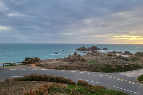 La Corbière Lighthouse stands beyond rugged rocks and a coastal road under a grey sky with a golden horizon.