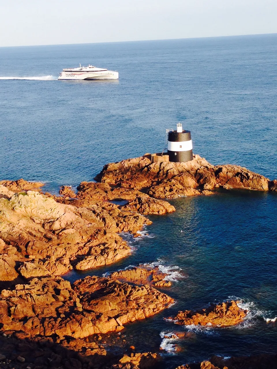 La Corbière's white lighthouse stands on warm-coloured coastal rocks with a ferry crossing the blue sea behind.
