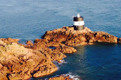 La Corbière's white lighthouse stands on warm-coloured coastal rocks with a ferry crossing the blue sea behind.