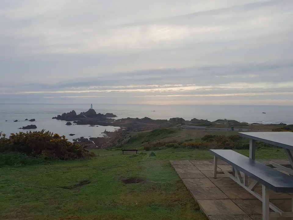 A picnic table overlooks grassy coastal slopes, rocky islets and La Corbière Lighthouse beneath a cloudy sky.