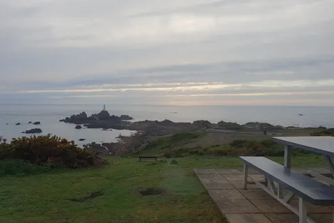A picnic table overlooks grassy coastal slopes, rocky islets and La Corbière Lighthouse beneath a cloudy sky.