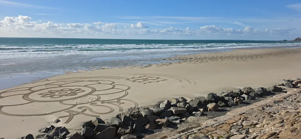 A large petal-shaped sand drawing stretches along the beach below stone sea defences and foamy surf.