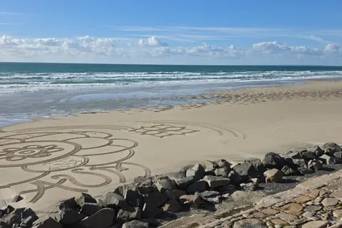 A large petal-shaped sand drawing stretches along the beach below stone sea defences and foamy surf.