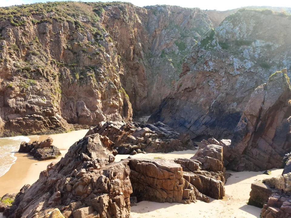 A rugged coastal rock face towers above sunlit boulders and a small patch of sand at Plémont.
