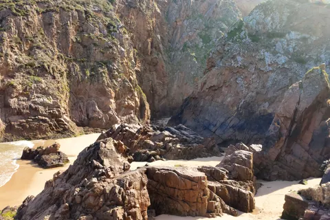 A rugged coastal rock face towers above sunlit boulders and a small patch of sand at Plémont.