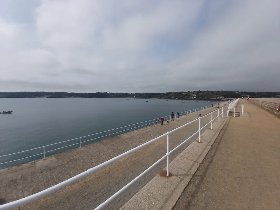 A broad stone walkway and white railings stretch into the distance beside calm water at St Catherine's Bay.