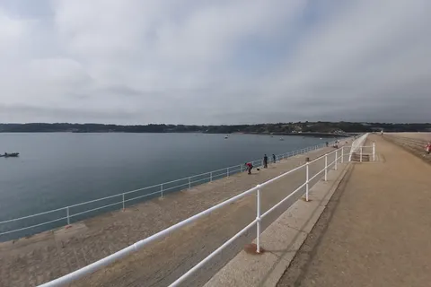 A broad stone walkway and white railings stretch into the distance beside calm water at St Catherine's Bay.