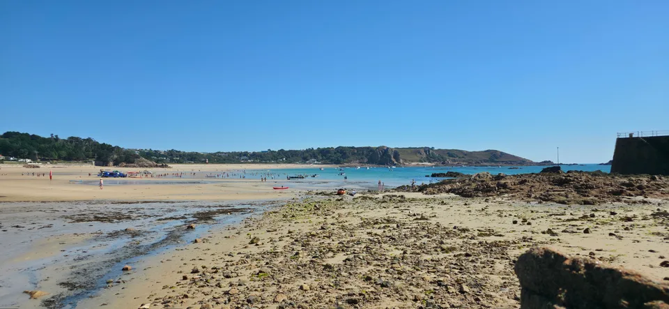 Seaweed-covered rocks and pale sand spread across the foreground of St Brelade's Bay below a clear blue sky.