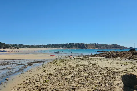 Seaweed-covered rocks and pale sand spread across the foreground of St Brelade's Bay below a clear blue sky.