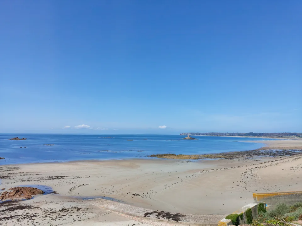 Pale sand and dark exposed rocks curve along the blue sea at St Ouen's Bay beneath a clear sky.
