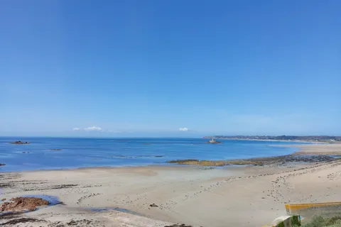 Pale sand and dark exposed rocks curve along the blue sea at St Ouen's Bay beneath a clear sky.