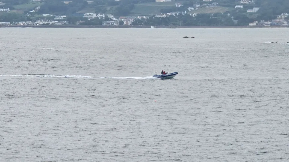A small motorboat leaves a thin white wake across grey-blue water in St Aubin's Bay.