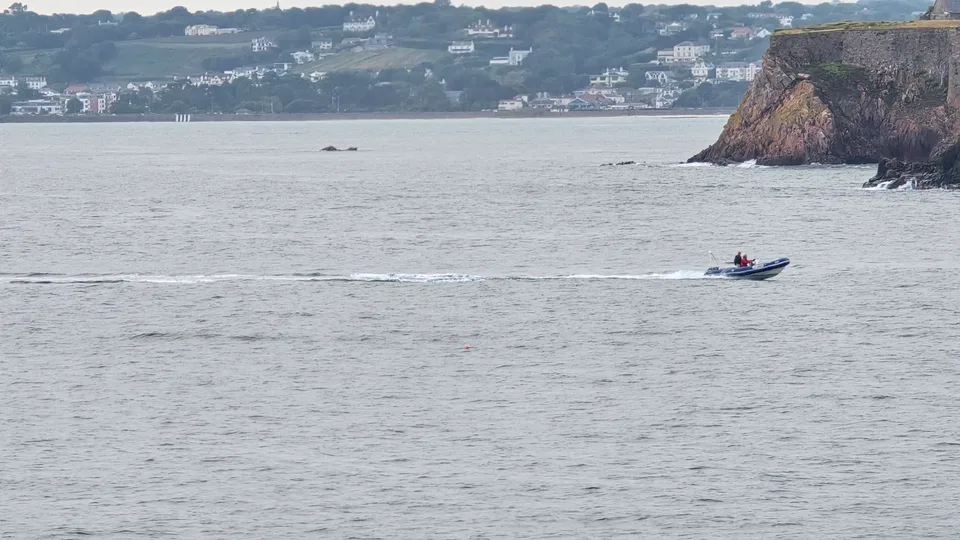 A motorboat passes beneath a rocky headland, with a long shoreline of hillside homes across St Aubin's Bay.