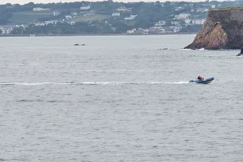 A motorboat passes beneath a rocky headland, with a long shoreline of hillside homes across St Aubin's Bay.