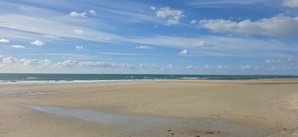 Broad pale sand and shallow breaking waves extend towards the horizon at St Ouen's Bay under blue sky.