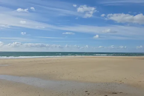 Broad pale sand and shallow breaking waves extend towards the horizon at St Ouen's Bay under blue sky.